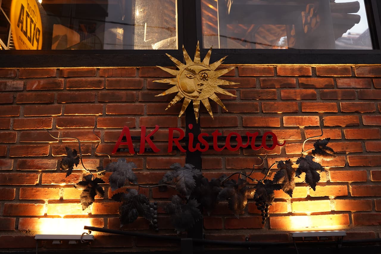 The AK Ristoro name in red script on an uplit brick wall, Ella — a brass sun and iron vine leaves around it
