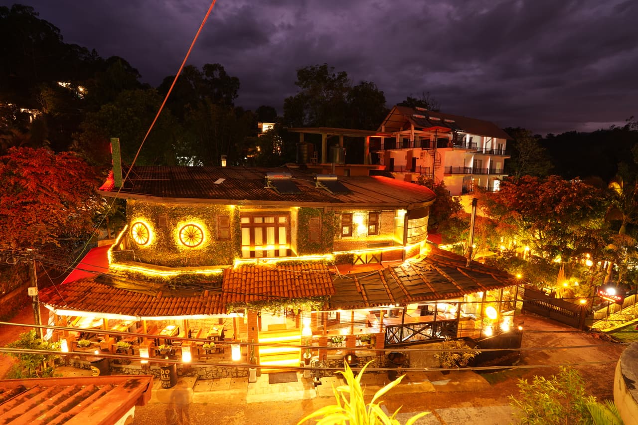 AK Ristoro, Ella, from above at blue hour — tiled roofs, lit wagon wheels and garden lights under a stormy sky