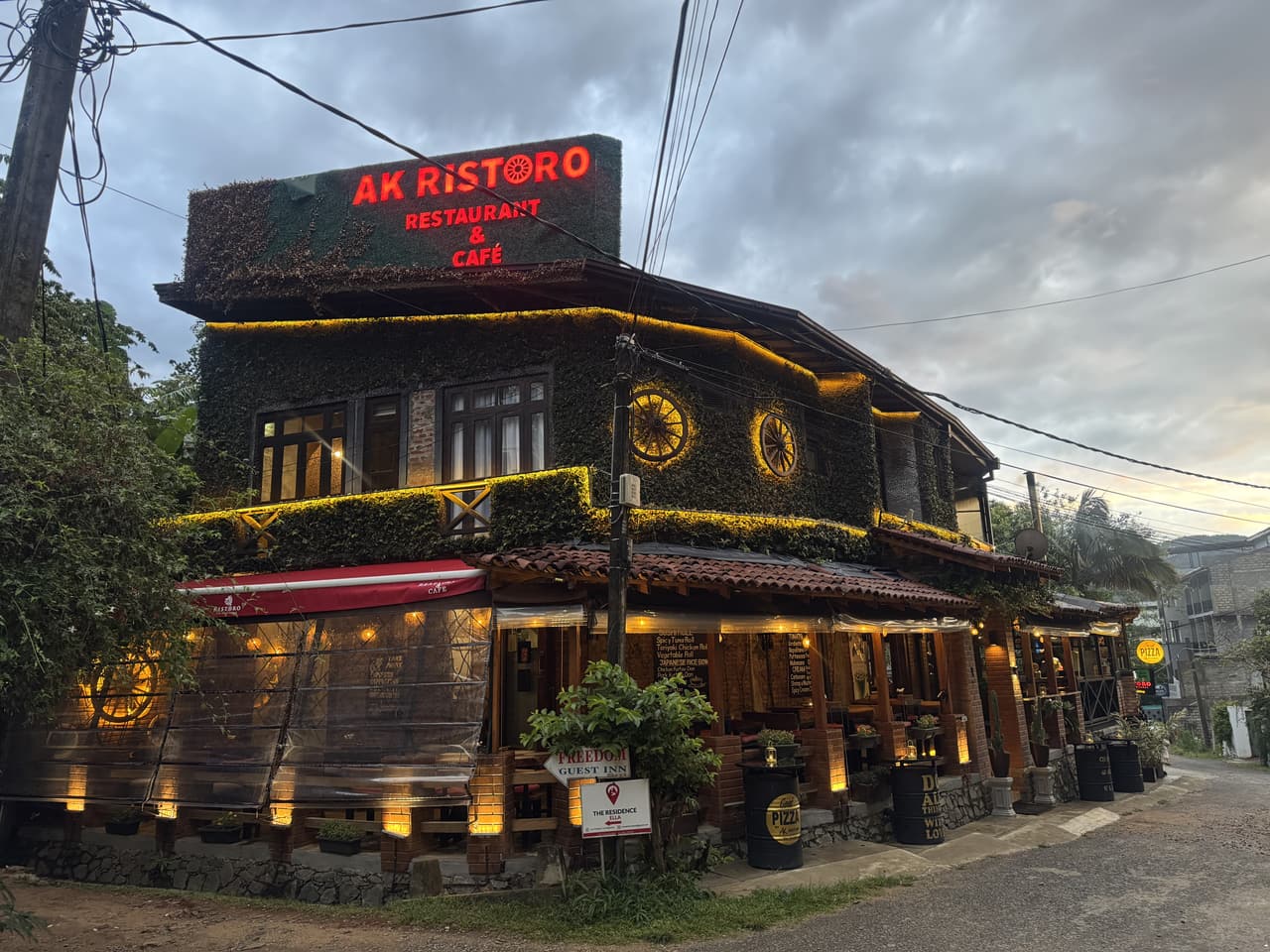 La esquina de AK Ristoro, Ella, al anochecer: el letrero rojo en lo alto del tejado, sobre las ruedas de carreta iluminadas