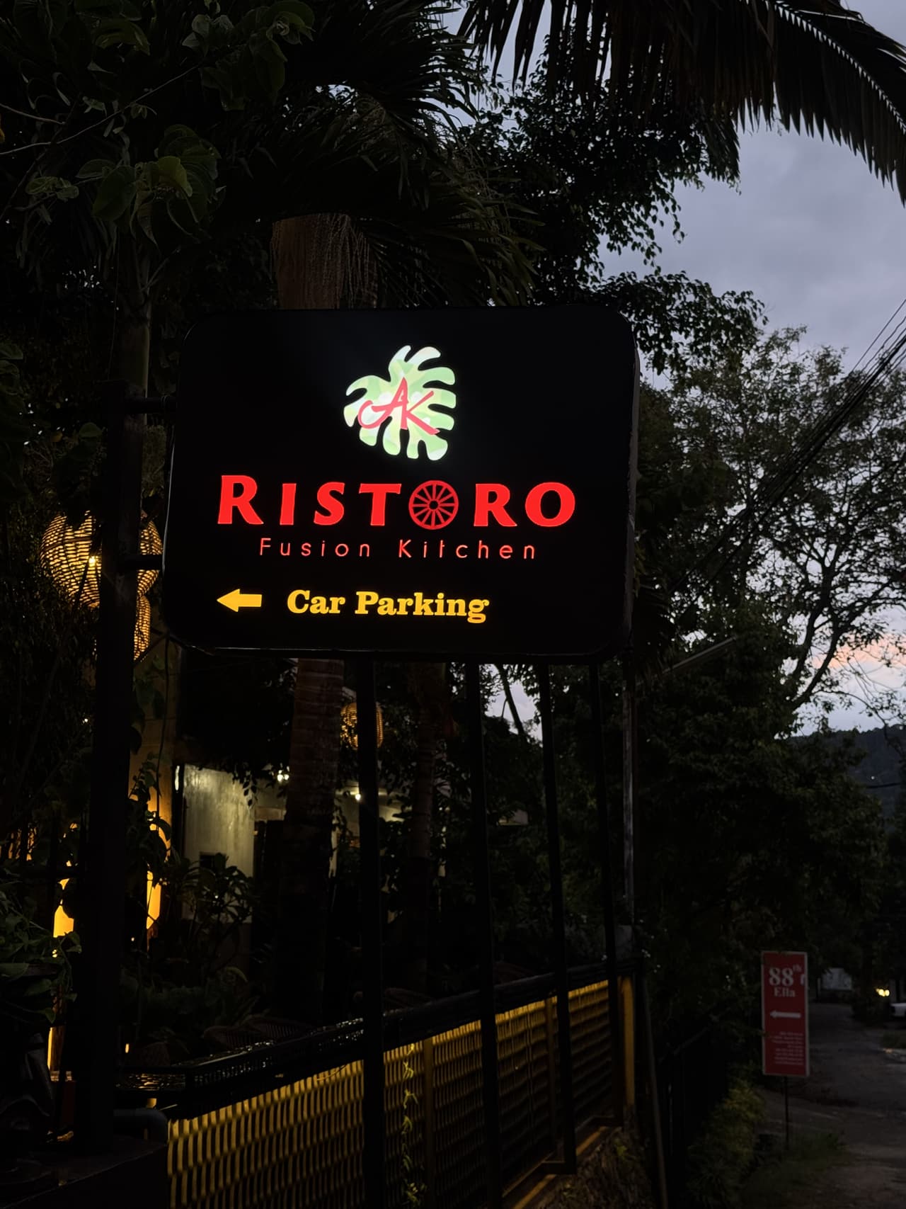 The illuminated Ristoro Fusion Kitchen sign on Passara Road, Ella, at dusk — the monstera leaf mark and an arrow to car parking