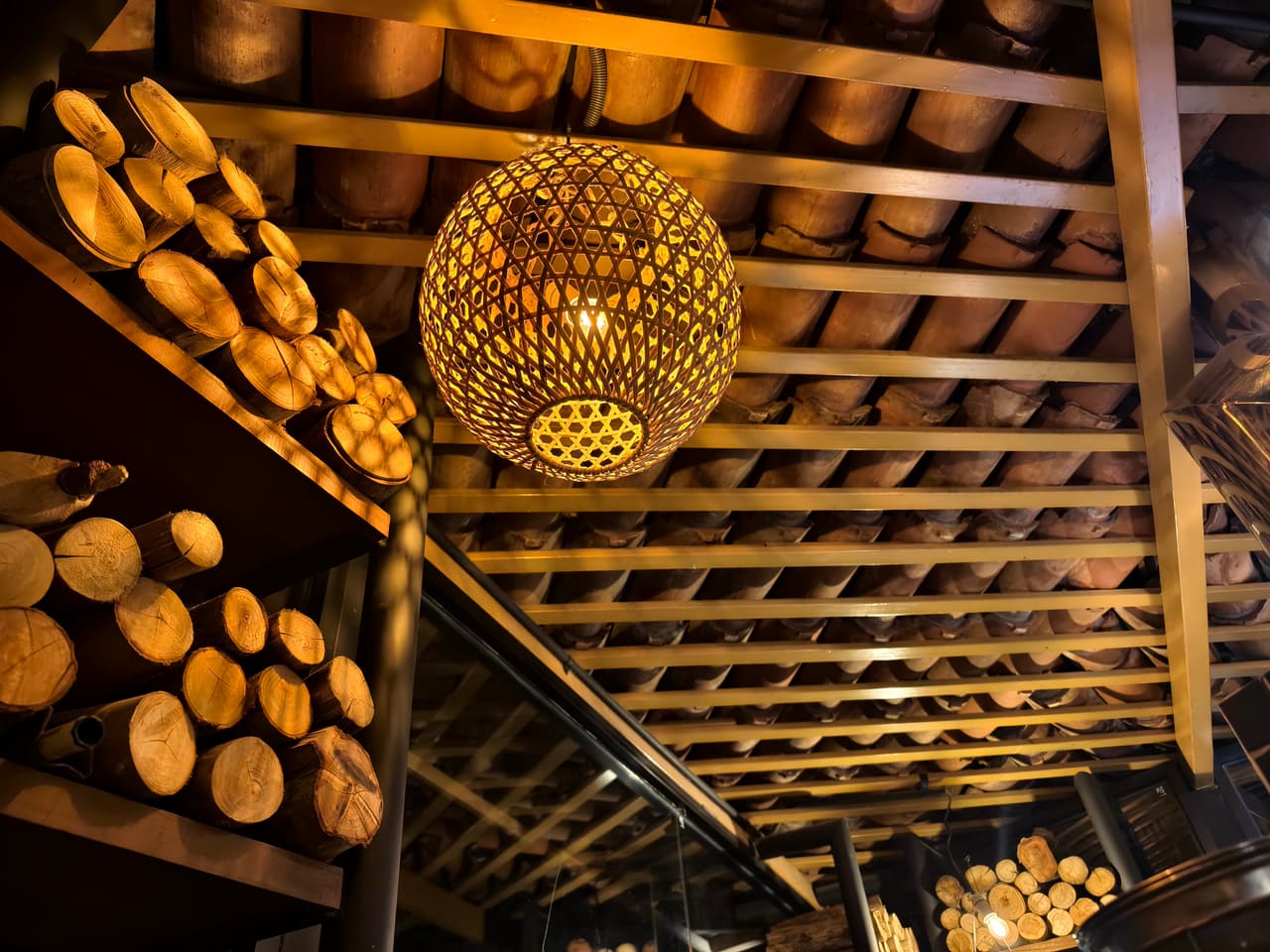 Firewood for the pizza oven stacked under the timber ceiling at AK Ristoro, Ella — a woven globe lamp glowing