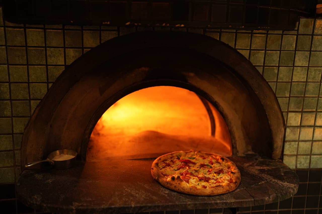 A pizza blistering at the mouth of the wood-fired oven at AK Ristoro, Ella — the dome glowing orange behind it