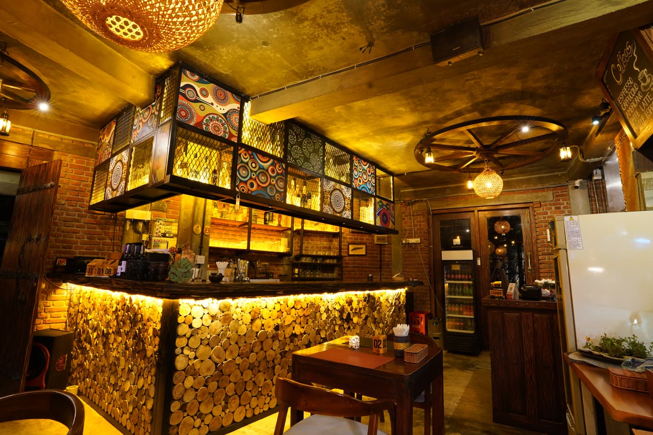 The bar at AK Ristoro, Ella — a counter faced with glowing log slices under patterned panels and a wagon-wheel chandelier